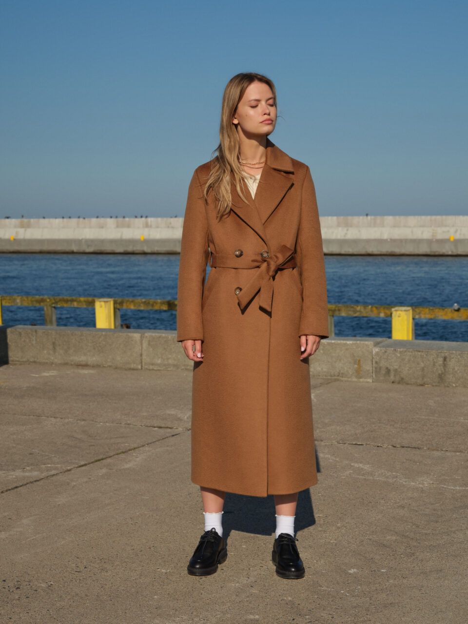 Camelowy długi płaszcz z wełny wielbłądziej o szlafrokowym kroju. Wiązany w pasie, zapinany na guziki. Uszyty z szlachetnej tkaniny o falistej strukturze, efekt zibellino.