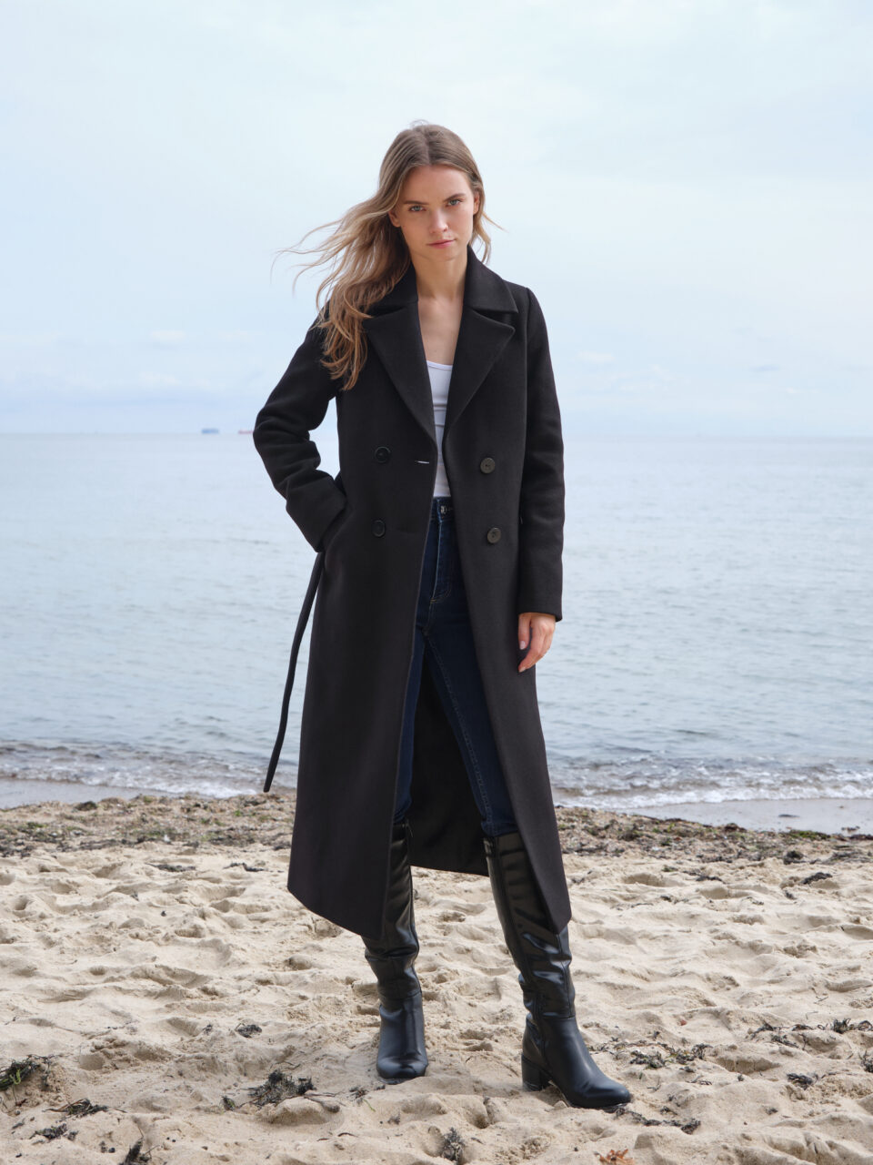 Czarny, dopasowany długi płaszcz zimowy z tkaniny z przewagą wełny. Zapinany dwurzędowo, wiązany. Minimalistyczny krój i bazowe kolory.