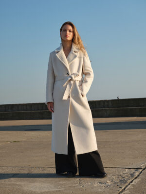 Długi zimowy kożuch w pięknym kremowym kolorze. Surowe wykonczenie, duży kołnierz, zapiany na guziki i wiązany w pasie. Ciepłe i stylowe okrycie w stylu teddy. Idealna baza dla wielowarstwowych, miejskich stylizacji.