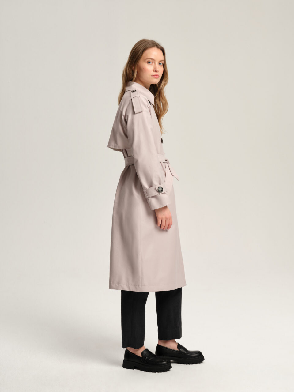 Długi luźny trencz damski w stylu oversize z raglanowymi rękawami wykończonymi ozdobnymi pagonami. Zapinany dwurzędowo na guziki i wiązany w pasie. Ozdobna peleryna na tyle.