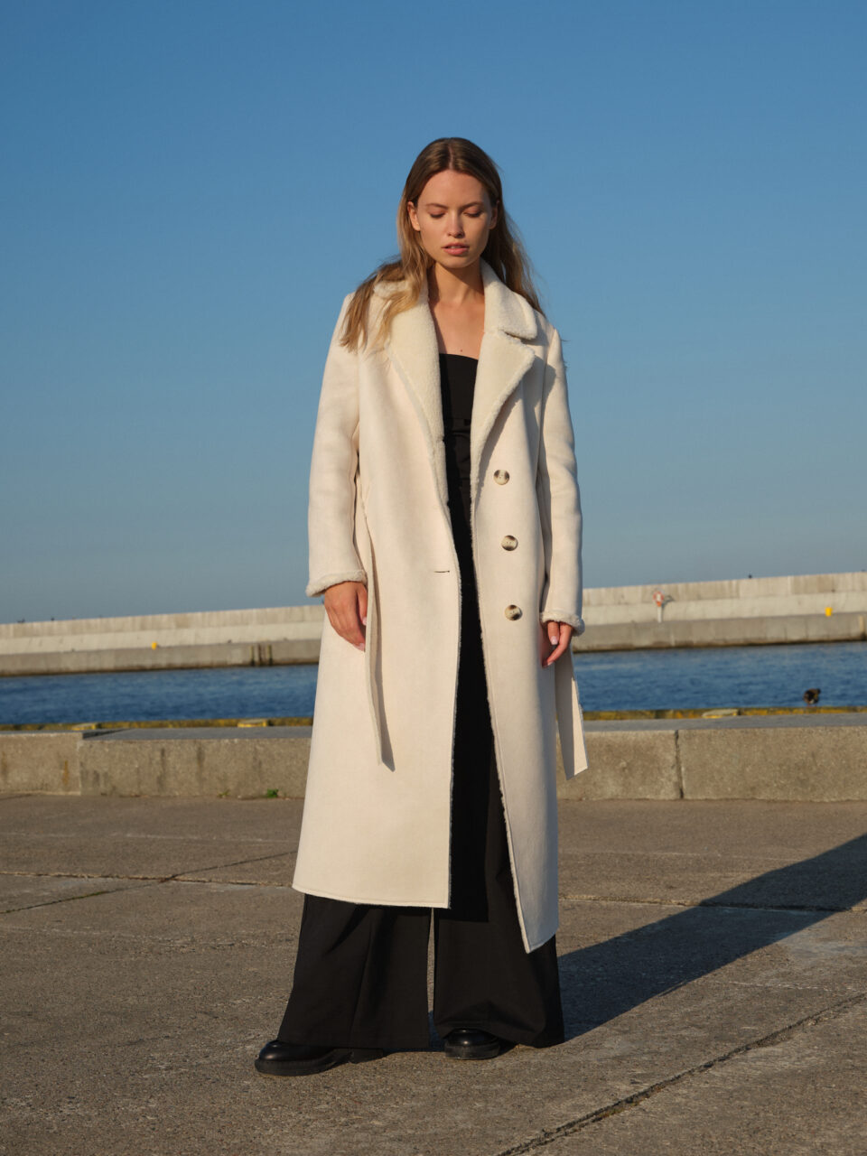 Kremowy długi kożuch zimowy z szerokim kołnierzem i futrzanym wykończeniem. Ciepły i stylowy. Wygodna linia pomieści wełniany sweter czy marynarkę. W stylu teddy.