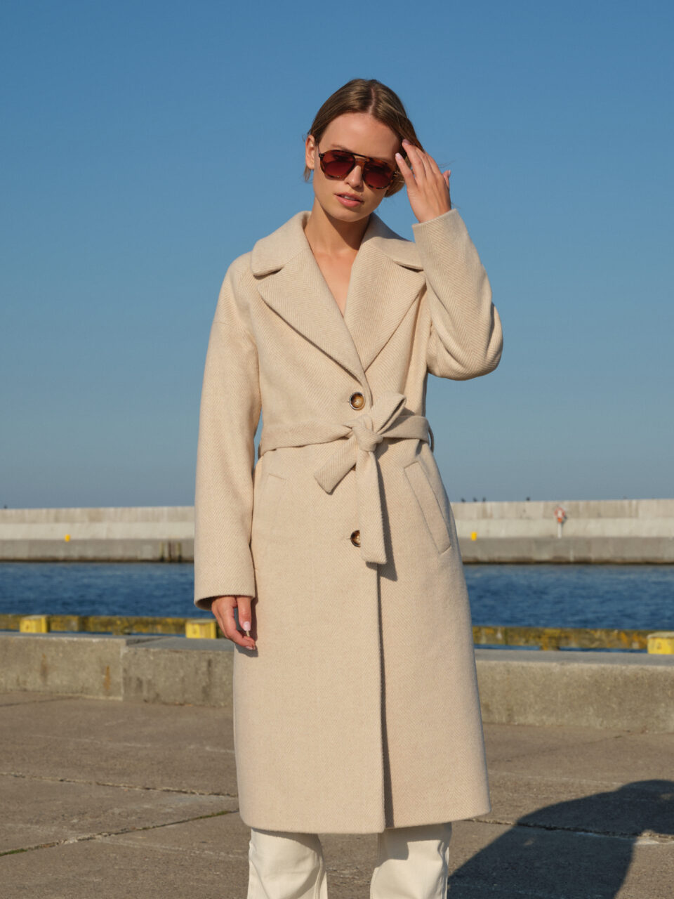 Zimowy, jednorzędowy płaszcz w beżową jodełkę z raglanowymi rękawami. Wiązany w pasie, zapinany na guziki. Klasyczny krój, świetny jakościowo materiał, ponadczasowy wzór jodełki. Utrzymany w trendzie Quiet Luxury.