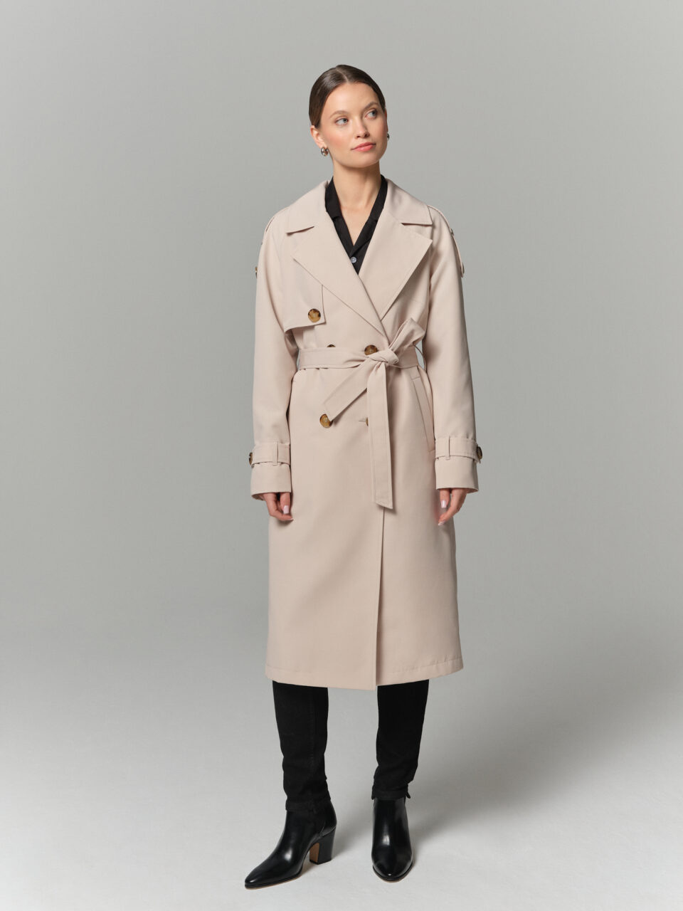 Długi trencz damski w chłodnym kremowym odcieniu. Zapinany dwurzędowo i wiązany. Ozdobne pagony i pasek na rękawach.